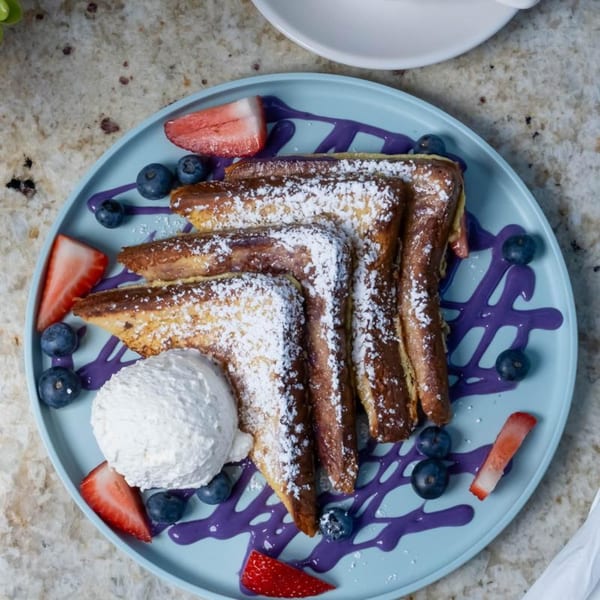 French toast with powdered sugar, whipped cream, blueberries, strawberries, and purple syrup on a blue plate.