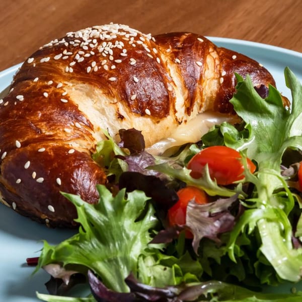 A pretzel croissant sandwich with cheese and meat, served with a side salad on a blue plate.