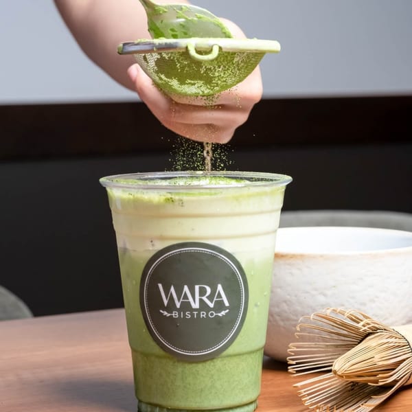 Hand sifting matcha powder over an iced matcha latte from Wara Bistro, with a whisk nearby.
