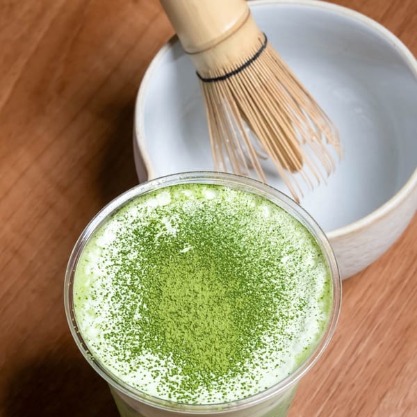 Matcha latte with green powder topping, next to a bamboo whisk and bowl on a wooden table.