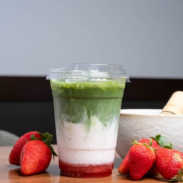 A layered strawberry matcha latte in a plastic cup with fresh strawberries on a wooden table.