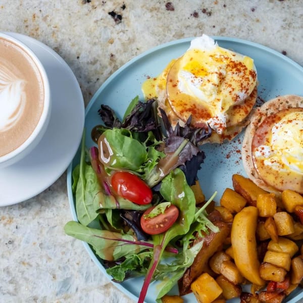 Eggs Benedict with a side salad, roasted potatoes, and a latte on a light blue plate.