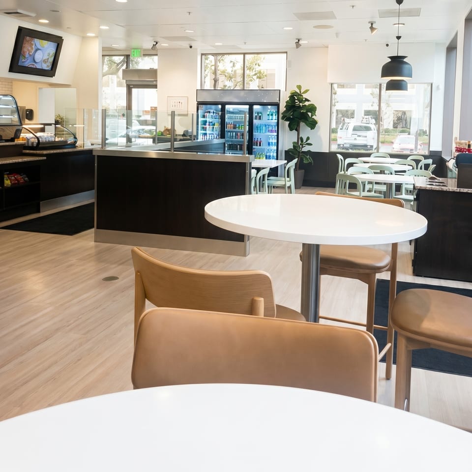 Modern cafe interior with light wood floors, white tables, and black counters with a beverage cooler.