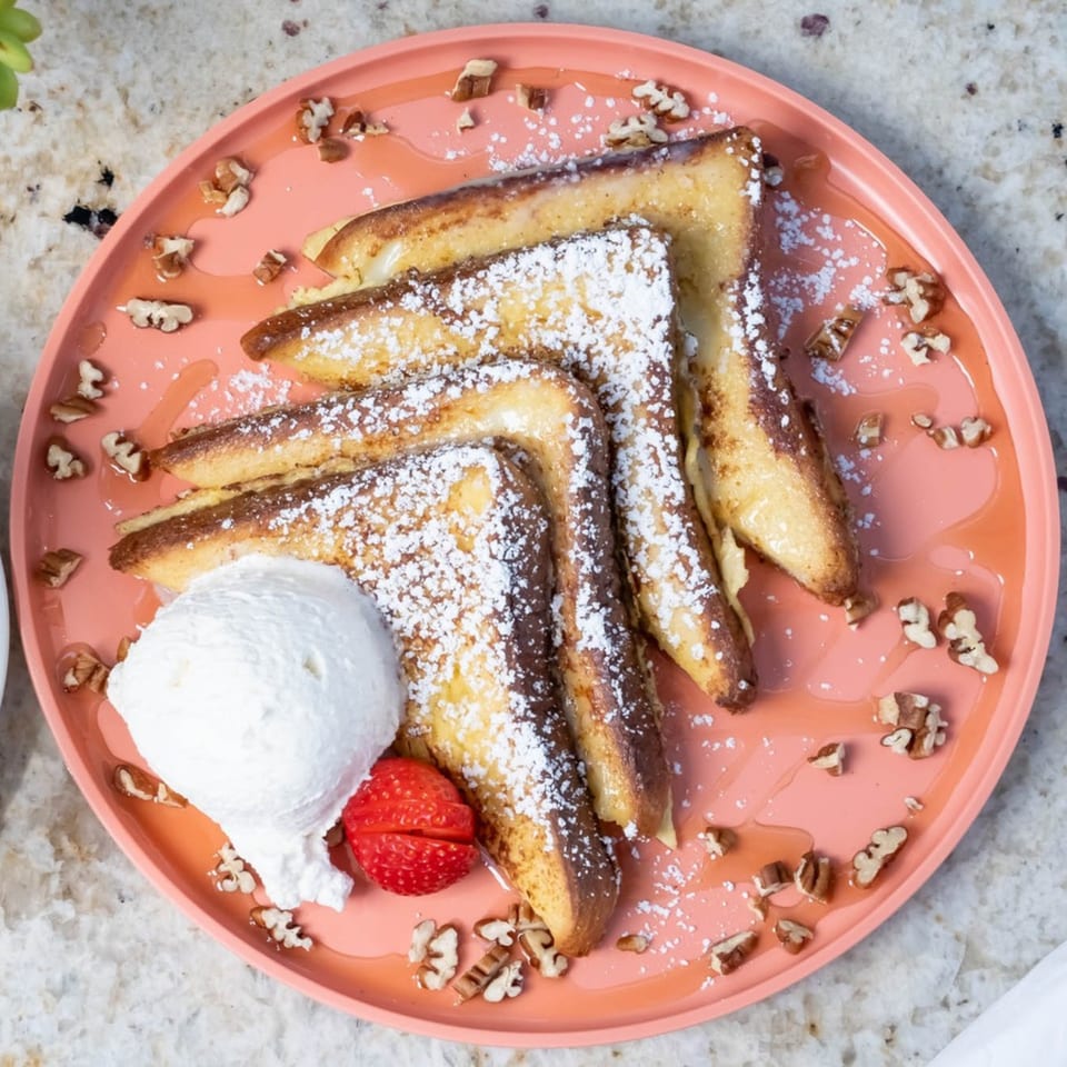 Golden French toast slices dusted with powdered sugar, topped with whipped cream, a strawberry, and pecans.
