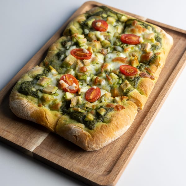 Rectangular pesto flatbread with chicken, melted mozzarella, and cherry tomato halves on a wooden board.