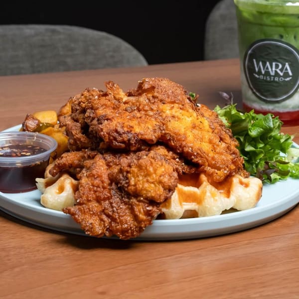 Crispy fried chicken and waffles with a side of sauce, potatoes, and salad greens.