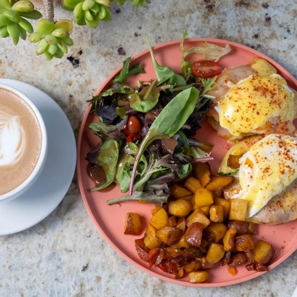 Eggs Benedict with home fries and a side salad on a pink plate, next to a latte.